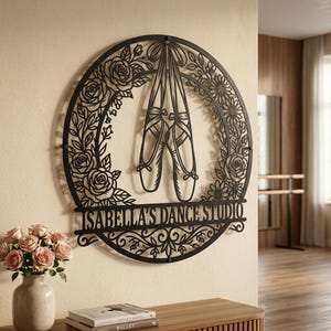 Könnte beinhalten: Ein rundes Wandbild aus schwarzem Metall mit einem ausgeschnittenen Design von Ballettschuhen, die von oben herabhängen, umgeben von Rosen- und Gänseblümchenmustern, mit dem Schriftzug ISABELLA'S DANCE STUDIO im unteren Bereich.