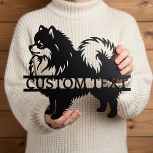 Pode incluir: Silhueta de metal preto de um cão Pomerânia com o texto "CUSTOM TEXT" recortado abaixo. O cão está em perfil, com uma cauda fofa e detalhes de pêlo. A placa é segurada em frente a um fundo de madeira.