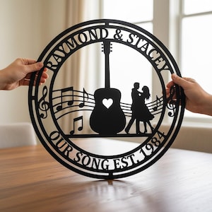 Könnte beinhalten: Schwarze Metall-Wandkunst mit Gitarre, Noten und tanzendem Paar. Der Text lautet "RAYMOND & STACEY" und "OUR SONG EST. 1984". Gehalten von zwei Händen. Ein dekoratives Element für das Zuhause.