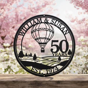 Könnte beinhalten: Schwarze Metall-Wandkunst mit einem Heißluftballon-Design, den Namen "WILLIAM & SUSAN" und der Zahl "50 YEARS". Das Kunstwerk enthält auch den Text "EST. 1974" und eine Landschaft.