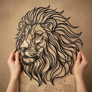 Op de afbeelding: Zwarte leeuwenkop wandkunst. De manen en gezichtskenmerken van de leeuw zijn ingewikkeld uit één stuk materiaal gesneden. Het kunstwerk wordt door handen omhoog gehouden, wat het gedetailleerde ontwerp en vakmanschap laat zien. De achtergrond is lichtbruin.