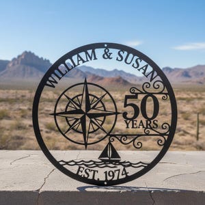 Könnte beinhalten: Schwarze Metall-Wandkunst mit einem Kompassrosen-Design, den Namen "WILLIAM & SUSAN" und dem Text "50 YEARS EST. 1974". Ein Segelboot ist auf den Wellen unter der Zahl 50 dargestellt. Die Kunst ist kreisförmig.