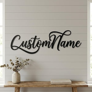 Könnte beinhalten: Schwarze Metall-Wandkunst mit dem Text "Custom Name" in Schreibschrift. Das Schild ist an einer hellgrauen Wand mit horizontalen Lamellen angebracht. Unter dem Schild befindet sich eine Holzbank mit einer Vase und einem Buch.