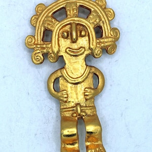 Puede incluir: Figura de metal dorado que representa una figura humana estilizada con un tocado. La figura tiene una cara sonriente, los brazos a los lados y pies detallados. El diseño recuerda al arte antiguo.