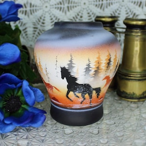 May include: A decorative ceramic vase with a black base, featuring a painted landscape with a running black horse silhouette, trees, and an orange and white sunset. A vintage brass telescope is next to the vase.