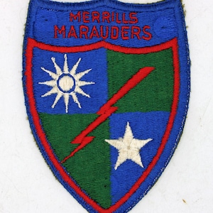 May include: A blue, green, and red embroidered patch with the text "MERRILLS MARAUDERS". The patch features a sunburst, a lightning bolt, and a star. The patch is shield-shaped.