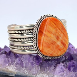 Pode incluir: Pulseira de punho de prata com uma grande pedra laranja oval. A pedra é fixada em uma moldura de prata com uma borda em forma de corda. A pulseira repousa sobre uma formação de cristal roxo.