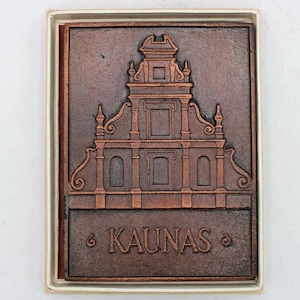 Puede incluir: Escultura en relieve de color cobre que representa un edificio, con la palabra "KAUNAS" en relieve en la parte inferior. La escultura está enmarcada en un marco blanco y marrón.