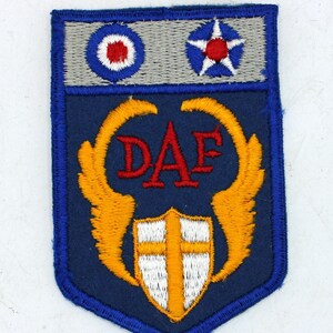May include: A blue and gray patch with embroidered details. The top section features a circular emblem and a star. Below, the patch displays the letters "DAF" in red, flanked by gold wings, and a white shield with a gold cross.