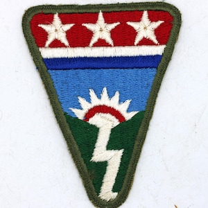 Puede incluir: Un parche triangular con una sección superior roja con tres estrellas blancas. Debajo, una franja azul y blanca, un campo azul con un sol rojo y blanco, colinas verdes y un rayo blanco. El parche está bordeado por un borde verde.