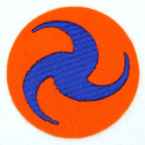 Op de afbeelding: Een ronde patch met een feloranje achtergrond en een blauw, driedelig wervelontwerp. De patch is gemaakt van een viltachtig materiaal, waarschijnlijk een embleem of insigne.