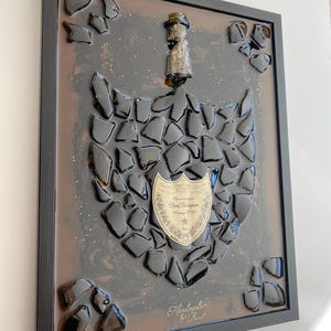 May include: A framed wall art piece featuring a flattened Dom Pérignon champagne bottle neck and label surrounded by black glass shards embedded in a dark, glittery resin background. The label reads Dom Pérignon Vintage 2015.