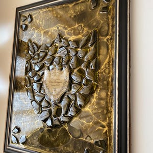 May include: A framed wall art piece featuring a central Dom Pérignon Vintage 2013 champagne label surrounded by a mosaic of dark green glass shards set against a swirling metallic gold and black resin background within a black wooden frame.