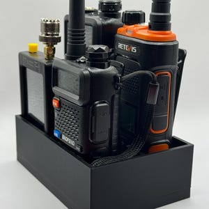 Könnte beinhalten: Ein schwarzer Funkgerätehalter mit mehreren Funkgeräten. Die Funkgeräte sind schwarz und orange, mit Antennen und verschiedenen Tasten und Displays. Der Halter ist ein einfaches, rechteckiges Design und bietet eine organisierte Aufbewahrung für Kommunikationsgeräte.