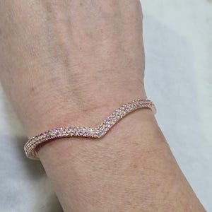 May include: Rose gold V-shaped bracelet. The bracelet is encrusted with small, iridescent crystals. The design features a gentle curve that tapers to a point in the center. The bracelet is worn on a wrist.