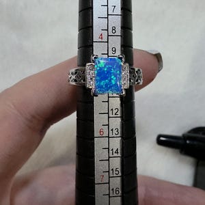 May include: A silver ring with a rectangular, blue opal center stone. The ring is set on a black ring sizer, with measurements visible. The opal has a vibrant, iridescent quality. The ring's band features decorative detailing with small, clear stones.