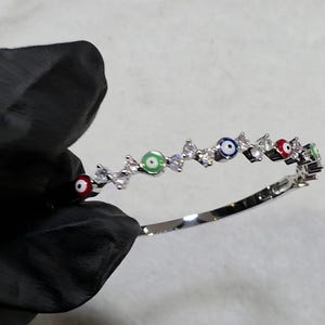 May include: A silver bracelet featuring a series of colorful evil eye charms alternating with clear, sparkling crystals. The bracelet is held against a white background, showcasing its intricate design and vibrant colors.