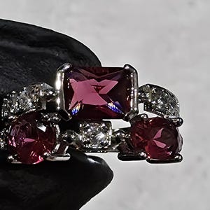 May include: A silver ring featuring a large, rectangular, magenta-colored gemstone as the centerpiece. The ring is accented with smaller, round, magenta stones and clear, sparkling accent stones. The ring is held by a gloved hand.