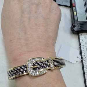 May include: Gold-toned bracelet with a buckle design. The buckle and a small rectangular accent are encrusted with clear crystals. The band is black and gold, resembling a woven pattern. A blank tag hangs nearby.
