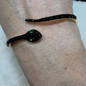Könnte beinhalten: Ein goldfarbenes Armreif-Armband in Schlangenform. Der Schlangenkörper ist mit schwarzen Kristallen besetzt, und der Kopf hat grüne Kristallaugen. Das Armband umschließt das Handgelenk und ist ein stilvolles Accessoire.