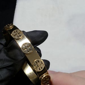 May include: Gold-tone metal bracelet featuring a series of circular embellishments with a four-leaf clover design. The bracelet is shown against a white background and is held by a person wearing a black glove.