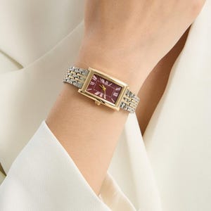 May include: A gold-toned rectangular wristwatch with a burgundy face and silver and gold-toned band. The watch is on a wrist, and the background is a white blazer.