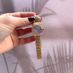 Puede incluir: Un reloj de pulsera dorado con esfera azul claro y visualización de fecha. El reloj tiene un bisel texturizado y una correa de eslabones. El reloj se sostiene en una mano sobre un fondo rosa.