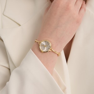 Könnte beinhalten: Eine goldfarbene Armbanduhr mit silbernem Zifferblatt und zartem Goldband. Die Uhr hat ein rundes Zifferblatt mit Stundenmarkierungen und Zeigern. Die Uhr wird am Handgelenk getragen, vor einem weißen Blazer.