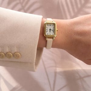 Pode incluir: Relógio de pulso retangular dourado com pulseira de couro creme. O mostrador do relógio tem algarismos romanos e ponteiros pretos. O relógio é usado no pulso, com a manga de um blazer bege ao fundo. A pulseira tem cerca de 1,2 cm de largura.