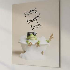 Könnte beinhalten: Ein witziger Druck, der einen grünen Frosch mit Sonnenbrille in einer weißen Badewanne mit Blasen zeigt. Der Text "Feeling froggin' fresh" steht über dem Frosch. Der Hintergrund ist hellbeige.
