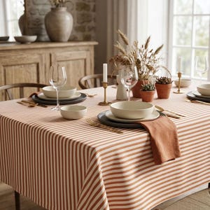 May include: A rectangular tablecloth featuring a pattern of vertical terracotta orange and white stripes. The table is set with white ceramic bowls, plates, clear glass wine glasses, gold-toned cutlery, and a folded terracotta orange linen napkin.