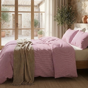 May include: A bed with a pink and white striped duvet cover and matching pillowcases. A beige throw blanket is draped over the bed. The room has a rustic aesthetic with a stone wall and large arched window.