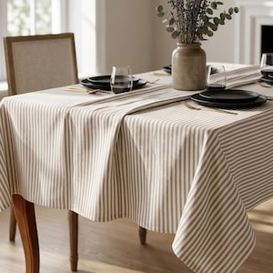 May include: A rectangular dining table covered with a beige and white vertical striped tablecloth. A matching table runner lies across the centre, topped with a ceramic vase containing dried lavender and eucalyptus stems, black plates, and glasses.