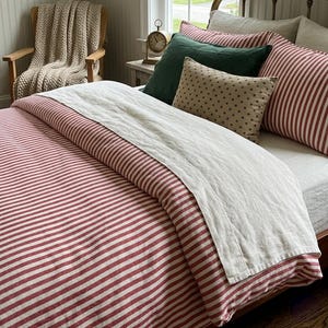 May include: A bedroom with a bed dressed in a red and white striped duvet and pillows. Additional green and star-patterned cushions are visible. A beige throw is draped over a wooden chair. A small clock sits on a bedside table.