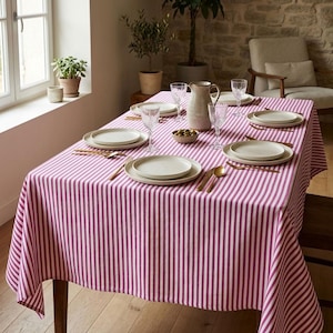 May include: A rectangular dining table covered with a magenta and white vertical striped tablecloth. The table is set with four white ceramic plates, clear textured glass goblets, gold-toned cutlery, and a central ceramic pitcher with olives.