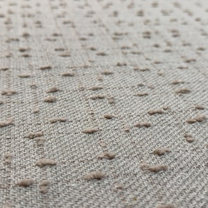 May include: A light beige fabric featuring a woven diagonal twill pattern and a series of small, raised, tufted dots in a matching beige tone distributed across the surface, creating a textured appearance with a soft, tactile quality.