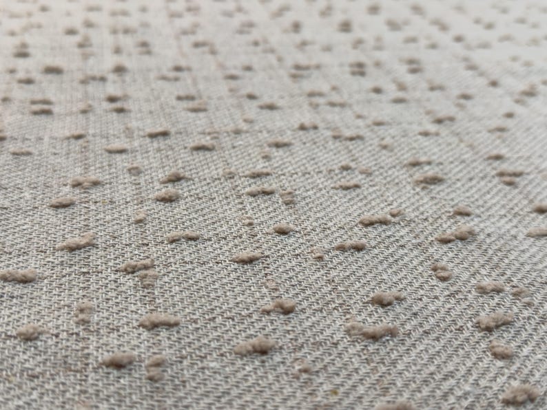 May include: A light beige fabric featuring a woven diagonal twill pattern and a series of small, raised, tufted dots in a matching beige tone distributed across the surface, creating a textured appearance with a soft, tactile quality.