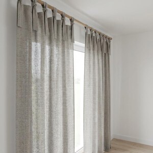 May include: A pair of light beige, semi-sheer curtains featuring a subtle, vertical speckled pattern in muted tones. The curtains are suspended from a wooden rod using fabric tie-top attachments, creating a soft, draped effect across the window.