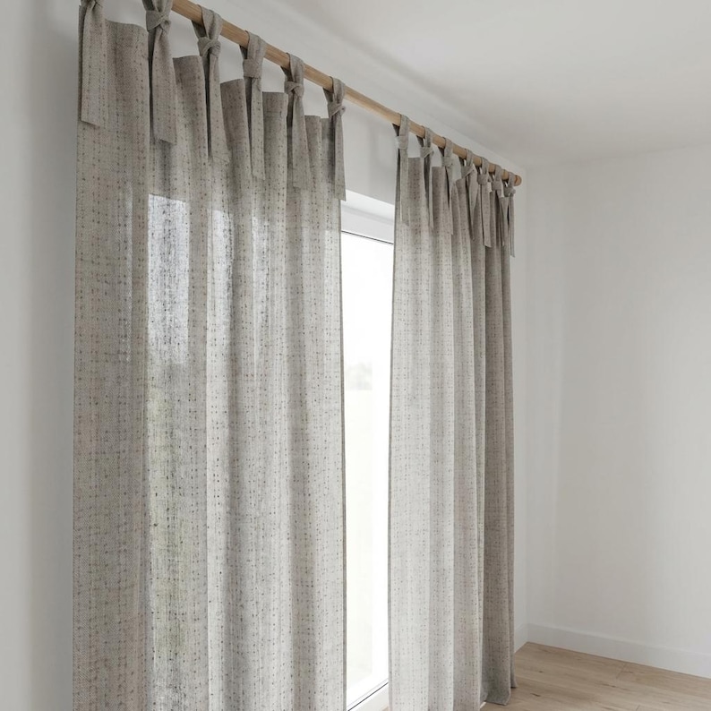 May include: A pair of light beige, semi-sheer curtains featuring a subtle, vertical speckled pattern in muted tones. The curtains are suspended from a wooden rod using fabric tie-top attachments, creating a soft, draped effect across the window.