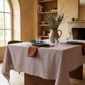May include: A rectangular tablecloth featuring a pattern of thin vertical brown and white stripes. The table is set with dark gray plates, clear glass tumblers, gold-toned cutlery, and a terracotta-colored napkin, with a ceramic pitcher.