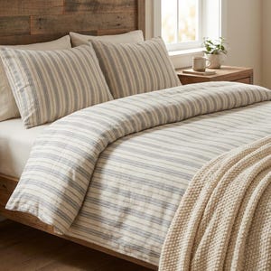 May include: A bed dressed with a cream duvet cover and pillowcases, both with blue vertical stripes. A beige knitted throw is draped over the bed. The wooden headboard and bedside table give a rustic feel to the bedroom.