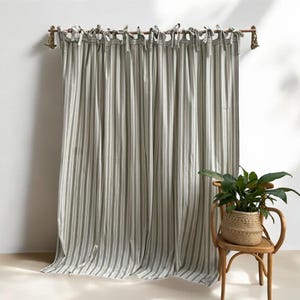 Op de afbeelding: Gestreepte gordijnpanelen in groen en wit, hangend aan een houten roede met decoratieve eindstukken. Het gordijn is gemaakt van een zachte stof en heeft een klassiek, verticaal streepjespatroon. Een geweven mand met een plant staat op een houten stoel.