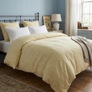 May include: A bedding set featuring a duvet cover with thin yellow and white horizontal stripes, two matching yellow and white striped pillow shams, two solid white rectangular pillows, and a folded light grey throw blanket on the bed.