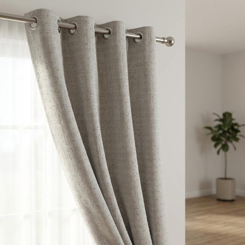May include: A light gray curtain panel with a textured, woven fabric appearance, featuring silver-toned metal grommets along the top edge. The curtain hangs from a matching silver-toned metal rod, creating soft, vertical folds across the fabric.