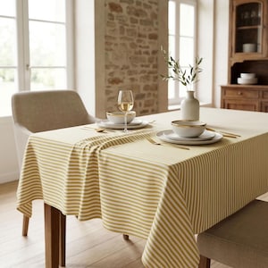 May include: A rectangular tablecloth featuring a pattern of horizontal mustard yellow and white stripes. The fabric drapes over a wooden dining table set with white bowls, gold-toned cutlery, a clear glass wine goblet, and a white ceramic vase.