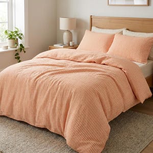 Könnte beinhalten: Ein Bett mit einer pfirsichfarbenen und weiß gestreiften Bettdecke und passenden Kissenbezügen. Die Bettwäsche befindet sich auf einem Holzrahmen, mit einer Lampe und einer kleinen Pflanze im Hintergrund. Der Raum hat eine neutrale Farbpalette.