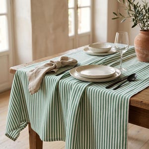 May include: A green and white striped tablecloth draped over a wooden table. The table setting includes a beige linen napkin, two stacked speckled ceramic plates, a clear glass wine goblet, a black spoon, and a black knife on the fabric.