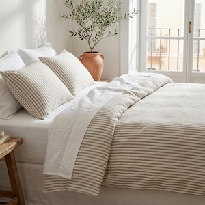 May include: A bed dressed with a striped duvet cover and pillowcases in beige and white. The bedding is on a white bed skirt. A terracotta pot with a plant is near a window, creating a serene bedroom scene.