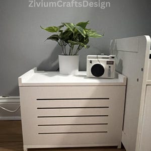 May include: A white storage chest with horizontal slats on the front, topped with a potted plant and a vintage-style camera. The chest is next to a white bed frame. The text "ZiviumCraftsDesign" is visible.