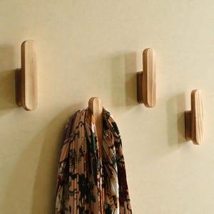 May include: Four wooden wall hooks with a curved design are mounted on a light-colored wall. One hook holds a patterned scarf with shades of pink, green, and brown. The hooks are made of light-colored wood.
